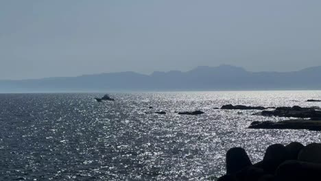 Silhouetten-Von-Felsen-Auf-Einem-Glitzernden-Meer-Mit-Booten,-Die-Unter-Einem-Klaren-Blauen-Himmel-Kreuzen