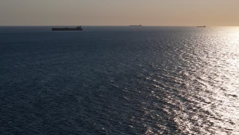 Tanker-Containerschiffe-Reihen-Sich-Am-Horizont-Auf,-In-Der-Ferne-Sind-Die-Silhouetten-Der-Boote-Zu-Sehen,-Während-Die-Drohne-Aufsteigt