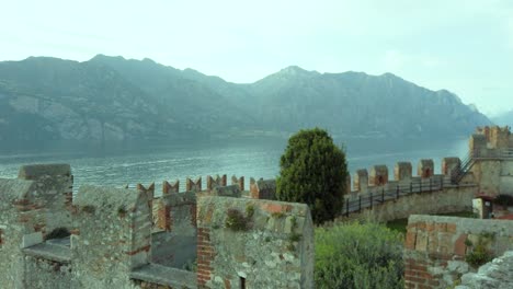 Mesmerizing-view-of-Lake-Garda-from-Malcesine,-featuring-the-parallax-effect,-with-a-backdrop-of-the-majestic-castle,-rocky-mountains,-and-lush-trees,-showcasing-the-rich-cultural-h