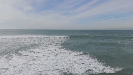 Toma-Aérea-De-Seguimiento-De-Un-Surfista-Surfeando-En-Una-Ola-Entre-Un-Grupo-De-Surfistas-En-El-Océano-Atlántico-Durante-La-Puesta-De-Sol-En-La-Playa-De-Caparica,-Portugal