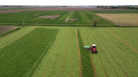 Farmer-sits-on-his-tractor-with-cutter-bar-and-mows-the-grass-of-his-pasture,-drone-flies-backwards-in-front-of-the-tractor
