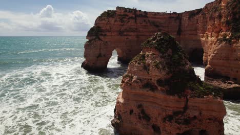 Luftaufnahme-Von-Zwei-Natürlichen-Bögen-Mit-Felsnadeln-Und-Wellen,-Die-Auf-Die-Malerischen-Klippen-Der-Estrada-Da-Caramujeira-In-Der-Algarve-In-Portugal-Krachen,-Per-4K-Drohne