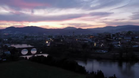 Twilight-descends-on-Barcelos,-showcasing-a-serene-river-and-historical-bridge-framed-by-a-pastel-sky