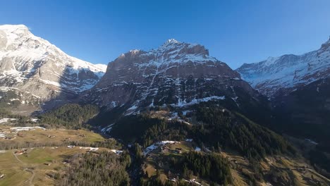 Steep-rugged-cliffs-of-Swiss-Alps-and-village-of-Grindelwald-in-shady-valley-far-below-snowy-mountain-peaks
