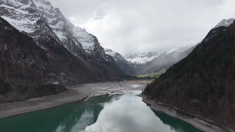 Aerial-view-of-Klöntalersee-with-a-reflective-turquoise-lake-nestled-between-snowy-mountain-peaks-and-steep-forested-slopes