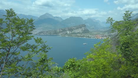 A-boat-glides-across-Lake-Garda-amidst-bushes,-under-a-blue-sky,-with-lush-greenery,-Italy