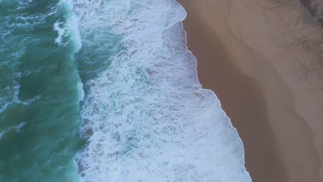 Aerial-view-tilting-over-waves-and-alone-women-walking-at-the-Magoito-Beach,-cloudy-sunset-in-Portugal