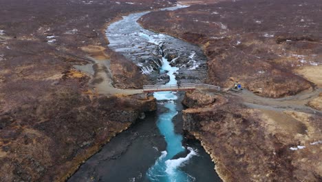 The-stunning-bruarfoss-waterfall-in-iceland-with-vibrant-blue-waters-and-rugged-terrain,-aerial-view