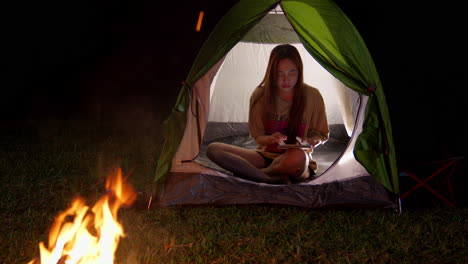 Asiatische-Frau-Beim-Camping-Im-Freien-Und-Chatten-In-Ihrem-Zelt-Mit-Einem-Modernen-Smartphone,-Das-Mit-5G-Verbunden-Ist,-Vor-Dem-Lagerfeuer