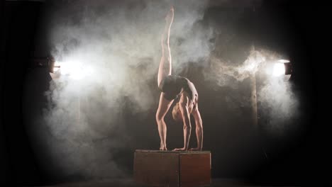 Acrobat-in-backbend-on-box-raise-leg-up,-stage-performance-in-smoky-spotlights