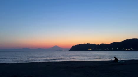 Una-Pareja-Sentada-En-Una-Playa-Al-Atardecer-Con-Vistas-A-Las-Montañas-Distantes-Y-Al-Vibrante-Cielo-Del-Atardecer