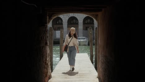 A-Woman-Is-Walking-Over-Wooden-Pier-Towards-Building-With-Arch-Passage-Along-Grand-Canal-In-Venice,-Italy