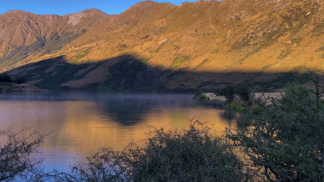 Sombra-De-Una-Montaña-Proyectada-A-Través-De-Un-Lago-Desde-El-Sol-De-La-Mañana,-Con-Niebla-Y-Un-Pescador