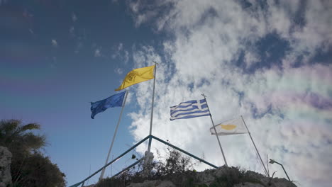 Flags-of-Cyprus,-Greece,-and-another-unidentifiable-flag-flying-high-on-a-pole-atop-a-rocky-outcrop-against-a-backdrop-of-blue-sky-and-clouds