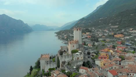 Aerial-view-of-Lake-Garda-and-Malcesine-Castle,-showcasing-the-charming-medieval-city-with-its-tiled-rooftops-nestled-amidst-the-breathtaking-Italian-mountains-and-serene-waters