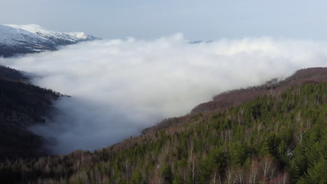 Luftaufnahme-Von-Wunderschönen-Bergtalhängen,-Die-Von-Schwebenden-Wolken-Bedeckt-Sind,-Schneebedeckter-Berggipfel-Am-Tag-In-Der-Ferne