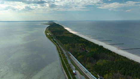 Aerial-view-capturing-the-unique-geography-of-Kuźnica-on-the-Hel-Peninsula,-showing-the-narrow-strip-of-land-with-a-railway-line-running-alongside-lush-greenery,-bordered-by-the-Baltic-Sea-and-bay