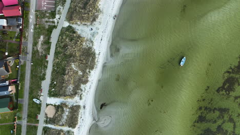 Aerial-shot-captures-a-lone-boat-floating-in-shallow-green-waters-near-a-white-sandy-beach-lined-with-natural-debris,-juxtaposed-against-vibrant-pink-buildings-and-a-bustling-street