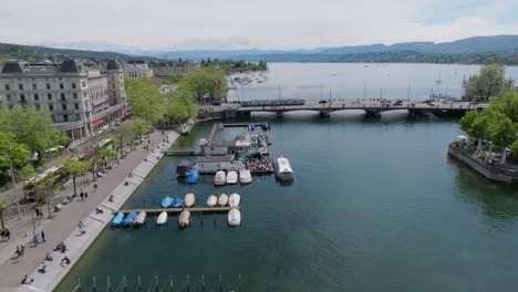 Forward-moving-drone-shot-showing-Zurich-City-bridge-and-Lake-Zurich-with-the-local-metro,-Swiss-flags-and-people-walking-across-the-bridge-with-the-lake-and-alps-visible-in-the-background
