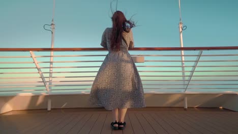 Woman-in-a-dress-on-the-deck-of-a-cruise-in-the-evening,-feeling-wind-and-looking-out-to-sea-back-shot-slow