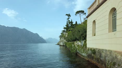 Explore-the-charm-of-Lake-Garda-from-Malcesine,-capturing-its-architectural-beauty-amid-lush-trees,-azure-waters,-and-scenic-skies