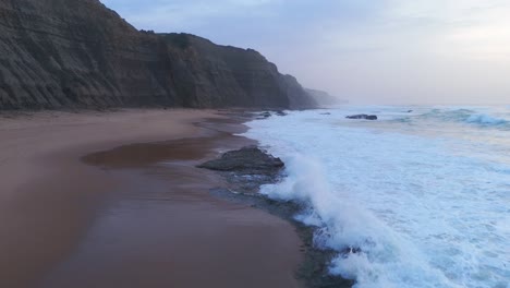 Aerial-view-tilting-over-waves-at-the-Magoito-Beach,-cloudy-sunset-in-Portugal
