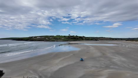 Inchidoney-sandy-beach-near-Clonakilty-in-the-morning-with-two-surfers-carrying-boards-across-with-gentle-waves-and-blue-sky,-Ireland