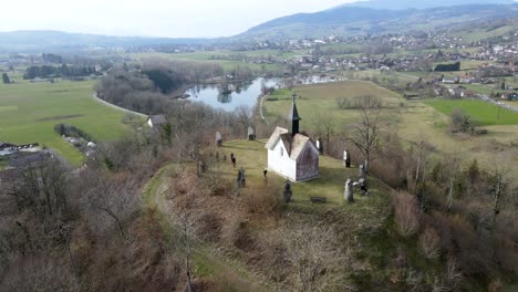 Surround-shot-of-a-white-church-on-top-of-a-hill-in-a-french-village-with-a-lake-and-mountains-in-the-background