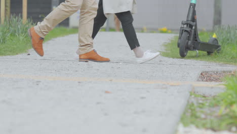 Lower-body-view-of-two-people-walking-on-a-concrete-pathway,-one-wearing-tan-pants-and-orange-shoes,-the-other-in-black-pants-and-white-shoes,-a-scooter-parked-to-the-side