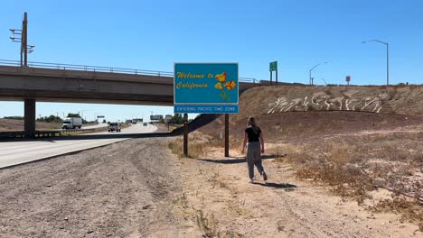 Happy-young-woman-traveler-walking-towards-Welcome-to-California-sign-at-USA-state-border