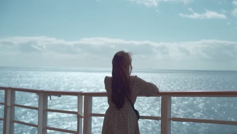 Woman-in-a-dress-on-the-deck-of-a-cruise-in-the-noon,-feeling-wind-and-looking-out-to-sea-back-shot-slow-long