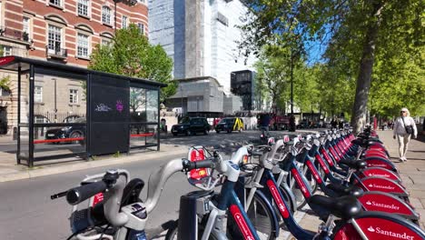 Reihe-Von-Leer-Geparkten-Santander-Leihfahrrädern-Stationiert-In-Westminster-Mit-Vorbeifahrenden-Radfahrern-Im-Hintergrund