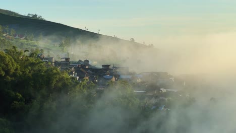 Foggy-village-on-mountain-side-in-Indonesia,-early-morning-sunrise,-aerial-view