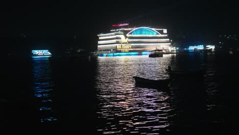 Nighttime-view-of-a-brightly-lit-casino-boat-on-water,-with-smaller-boats-nearby,-reflecting-vibrant-lights-on-the-surface
