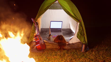young-girl-chatting-with-laptop-inside-her-tent-while-camping-outdoor-at-night-in-front-at-bonfire
