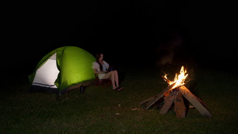 digital-nomad-working-remotely-concept,-asiatic-young-woman-sitting-with-laptop-outside-her-tent-in-front-of-bonfire-at-night