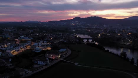 Aerial-view-of-Barcelos-at-dusk-with-lit-streets-and-ancient-bridge-over-river,-against-a-vibrant-sunset-sky