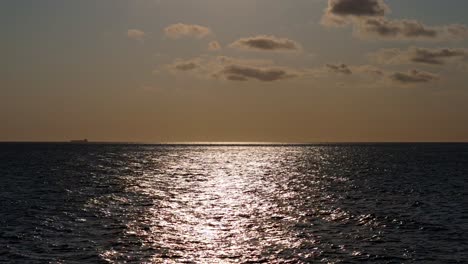 Tilt-down-from-sun-high-in-sky-with-tanker-on-horizon-as-sunlight-glistens-on-dark-water
