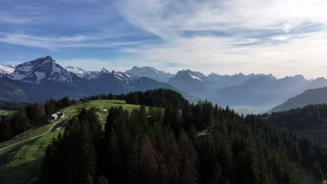 Aerial-view-of-lush-green-hills-with-dense-forests-in-the-foreground-and-snowy-mountain-peaks-in-the-distance-under-a-cloudy-sky,-Amden-Arvenbüel-Schweiz