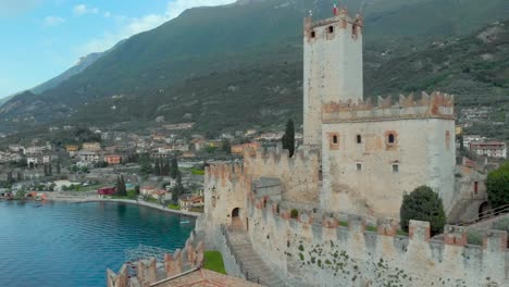 Vista-Aérea-Que-Revela-El-Castillo-De-Malcesine-Y-Su-Pueblo-Medieval-Enclavado-Entre-Agua,-Cielo-Y-Montañas.