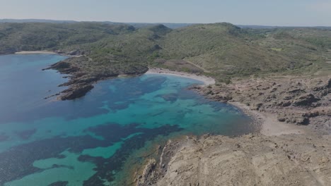 Drones-Aéreos-Sobrevuelan-La-Playa-Virgen-De-Cala-Sa-Torreta-En-La-Costa-Española-De-La-Isla-De-Menorca,-Paisajes-Naturales-De-Viaje,-Mar-Azul,-Colinas-Verdes-Alrededor-De-La-Costa-Arenosa-Y-Rocosa