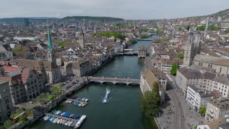 Fast-forward-moving-drone-shot-of-Zurich-city,-showing-old-town-architecture,-clock-towers-and-cities-skyline-with-river-in-the-foreground