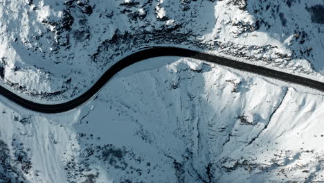 A-winding-road-cutting-through-a-snow-covered-mountain-landscape,-captured-in-bright-daylight,-aerial-view