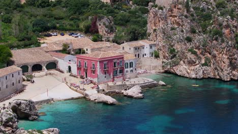 Feste-Luftaufnahme-Eines-Wunderschönen-Rosafarbenen-Gebäudes-An-Der-Mittelmeerküste-Mit-Blauem-Wasser-Und-Felsigen-Klippen