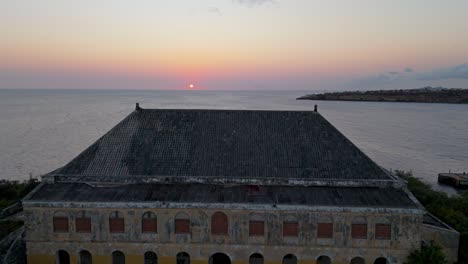 Encanto-Del-Viejo-Mundo-Edificio-Abandonado-Amarillo-Con-Vistas-Al-Océano-Que-Revela-El-Sol-Poniente-En-El-Horizonte,-Drone