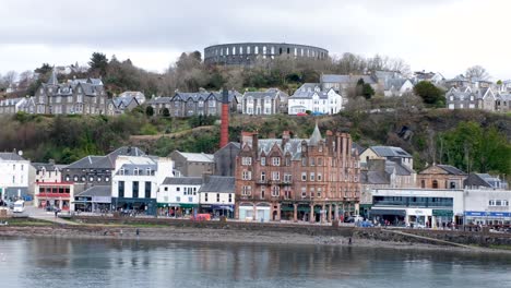 Scenic-landscape-view-of-Oban-town-with-people-and-traffic-along-the-harbour-waterfront-and-McCaig's-Tower-on-top-of-hills-in-Western-Scotland-UK