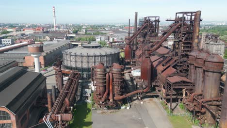 Aerial-view-of-rusted-industrial-plant,-juxtaposed-against-a-modern-cityscape-under-a-clear-sky