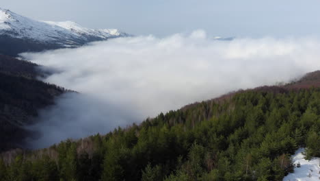 Luftaufnahme-Von-Wunderschönen-Berghängen-Mit-Bäumen,-Die-Von-Schwebenden-Wolken-Bedeckt-Sind,-Schneebedeckter-Berggipfel-Am-Tag-In-Der-Ferne