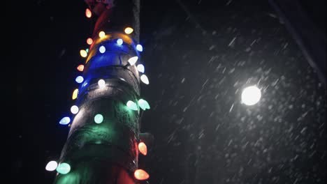 Winter-snowfall-at-night-snowing-on-Christmas-lights-on-city-street-in-street-light