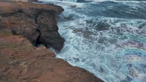 Las-Fuertes-Olas-Rompen-Contra-Los-Acantilados-Rocosos-Con-Una-Formación-Similar-A-Una-Cueva-Visible,-Creando-Una-Espectacular-Escena-Natural-A-Lo-Largo-De-La-Costa.